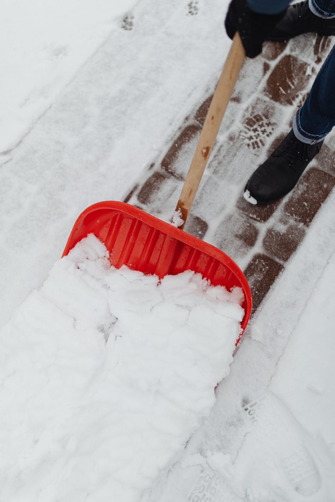 Gebäudereiniger schiebt im Winterdienst mit einer roten Schneeschaufel Schnee vom Pflasterweg